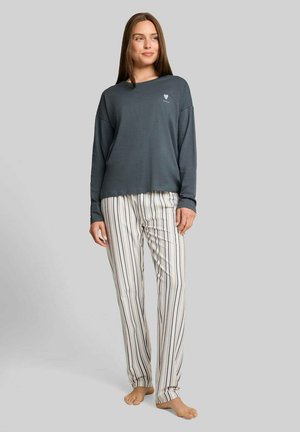 Junge Frau steht barfuß, trägt ein lockeres dunkles Langarmshirt und helle gestreifte Pyjamahosen vor einem schlichten grauen Hintergrund.