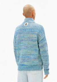 Gebreide trui in verschillende tinten blauw en groen met een hoge kraag, geribbelde textuur en een label met een gestileerd logo aan de achterkant.