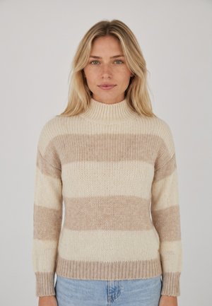 Femme blonde portant un pull en maille beige et crème rayé avec un col montant et un jean bleu clair sur un fond uni.