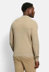 Beige gestrickter Pullover mit Kragen, gerippten Bündchen und Saum. Besitzt ein strukturiertes Muster und eine lockere Passform. Getragen mit beigen Hosen.