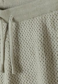 Tissu tricoté beige avec une ceinture côtelée et un cordon de serrage, présentant une texture en maille ouverte à motif.