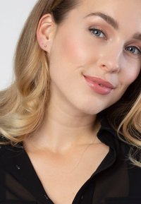 Gouden studoorbellen bij een persoon met golvend blond haar en een delicate gouden ketting. De top is zwart met een subtiele textuur.