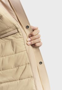 Beige pufferjack geïnterior met een gladde stofafwerking, zichtbare gewatteerde textuur, een ritssluiting en een detail van een zijzak.