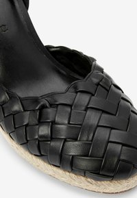 Espadrille noire en cuir tressé avec une surface texturée et une semelle en jute naturel. Présente des bords festonnés pour une finition douce.