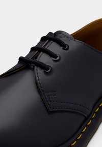 Gros plan d'une chaussure en cuir noire à lacets avec trois œillets et une couture jaune le long du bord de la semelle.