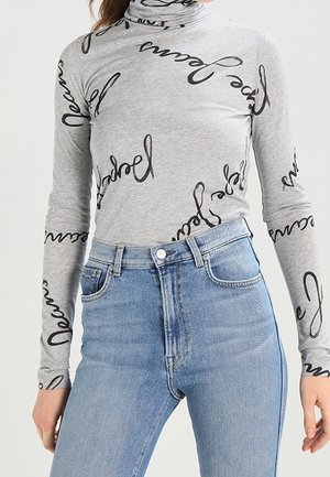Col roulé gris à manches longues avec motif de texte en cursive noire, associé à un jean en denim bleu clair taille haute porté par une personne debout.
