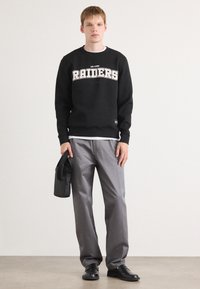 Sweatshirt preto com gráfico e logótipo "RAIDERS", calças cinzentas e sapatos pretos. Acessórios incluem uma pequena mala preta. Ajuste casual, design simples.