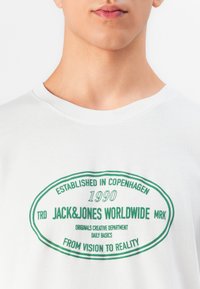 Jaunas žmogus, vilkintis baltą marškinėlį su žaliu ovaliu logotipu, ant kurio yra užrašas "Jack&Jones Worldwide, įsteigta Kopenhagoje 1990, nuo vizijos iki realybės."
