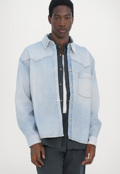 Lyseblå denimjakke med knappefront, spiss krage, brystlomme og avslappet passform. Laget over en svart skjorte med knapper.