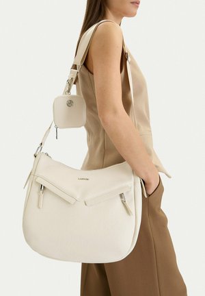 Mujer con top beige sin mangas y pantalones marrones, llevando un bolso grande de hombro color crema con una pequeña bolsa a juego adjunta.