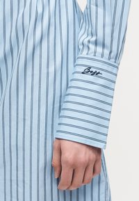 Φωτεινό μπλε μανίκι πουκάμισου με λεπτές κάθετες ναυτικές ρίγες και μαύρη κεντημένη υπογραφή στη μανσέτα, εμφανιζόμενο με χαλαρό χέρι.