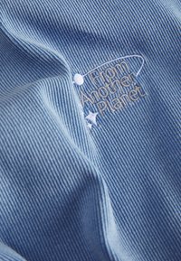 Světle modrá žebrovaná látka s bílou výšivkou s textem "Z jiného světa" a malými hvězdnými akcenty. Měkká textura.