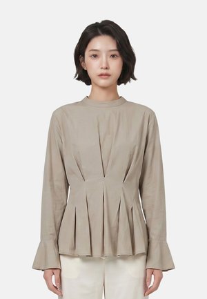 Vrouw met kort donker haar draagt een beige blouse met lange mouwen, plooien en uitlopende manchetten, gecombineerd met lichtgekleurde broek op een witte achtergrond.