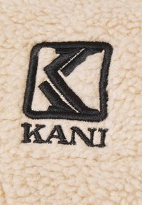Tissu texturé de couleur crème avec un logo noir brodé affichant un motif géométrique et le mot "KANI" en dessous.