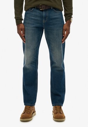 Hombre con vaqueros azules gastados, cinturón de cuero marrón, botas marrones con cordones, suéter verde oliva y reloj de pulsera dorado, de pie con las manos a los lados.