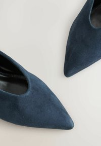 Zapatos de tacón alto de gamuza azul marino con punta afilada y detalles recortados en los laterales, que destacan por su textura suave y diseño elegante.