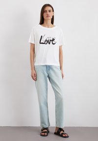 Weißes Baumwoll-T-Shirt mit schwarzem "Love"-Grafik, lässiger Schnitt. Kombiniert mit hellblauen Jeans und schwarzen Sandalen.
