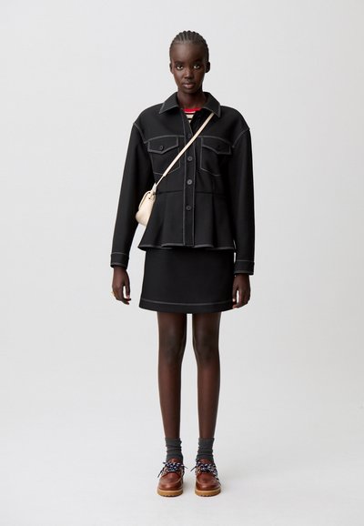 Giacca di jeans nera con cuciture bianche, abbinata a una gonna nera. La modella indossa calzini grigi e scarpe marroni con accenti blu.