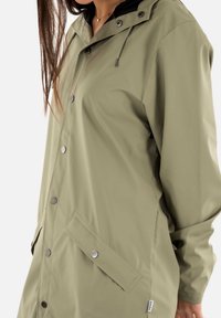 Chaqueta impermeable verde con capucha, que cuenta con un cierre frontal a presión, bolsillos laterales y una textura suave. Cuerda ajustable en la capucha.