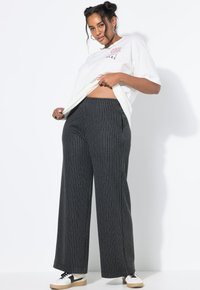 Femme soulevant un T-shirt blanc avec le texte "Cool Vibes Here", portant un pantalon large à rayures grises et des baskets blanches, se tenant devant un fond blanc.