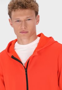 Červená mikina s kapucí na zip, podšitá fleece, s černým zipem, nošená přes bílý tričko s kulatým výstřihem. Hladká textura, přiléhavý střih.