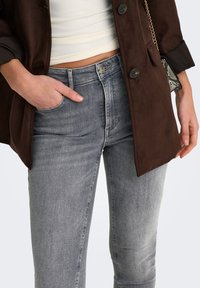 Bruine suède jas met een knoopsluiting aan de voorkant, gecombineerd met grijze slim-fit jeans met klassieke zakken en een vervaagde wassing.