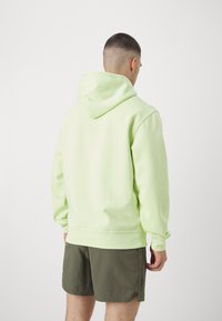Felpa con cappuccio verde chiaro, dal taglio morbido, con polsini e orlo a coste, dotata di un cappuccio ampio e dettagli minimi. Abbinata a pantaloni corti scuri.