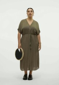 Olive-green, loose-fitting dress with a V-neck and short sleeves, cinched at the waist with a drawstring. Black woven bag and chunky shoes.
