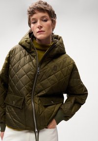 Olivgrüne Steppjacke mit Reißverschluss, Kapuze und zwei Vorderaschen. Strukturierter Stoff und gerippte Bündchen heben ihr Design hervor.