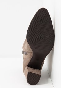 Gros plan sur la semelle et le talon d'une bottine en daim taupe avec fermeture éclair latérale, montrant une semelle extérieure en caoutchouc noir texturé et un talon bloc.