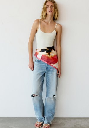 Jeune femme aux cheveux blonds portant un caraco blanc, une grande écharpe fleurie nouée autour de la taille sur un jean bleu clair déchiré et des sandales.