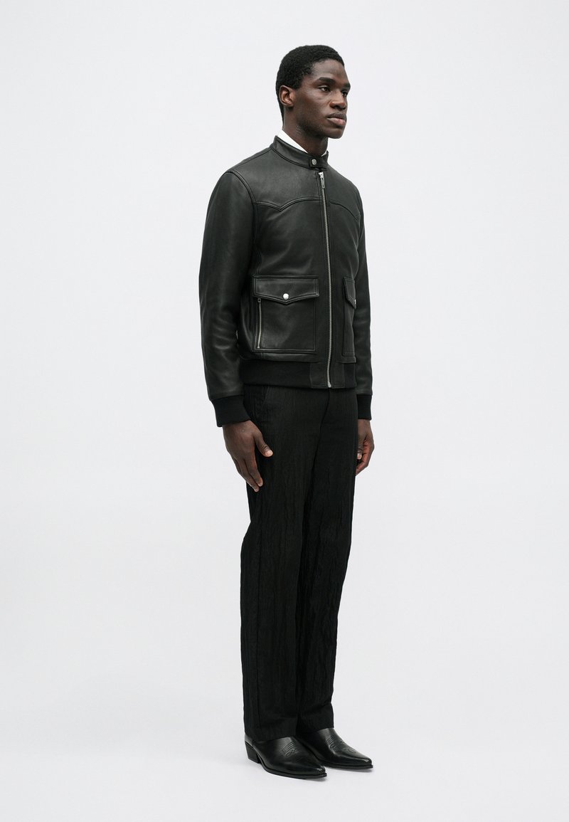 Black leather bomber jacket with two front patch pockets, ribbed cuffs, and zip closure. Paired with textured black pants and black boots.