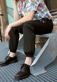 Mustat joustavat hihansuutet housut yhdistettynä kukkakuosisella napitettavalla paidalla. Kengät ovat mustaa nahkaa nauhoilla, istuen metallisilla portailla.