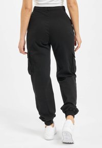 Personne portant un pantalon de jogging cargo noir avec des poignets élastiques et des baskets blanches, se tenant devant un fond blanc uni.