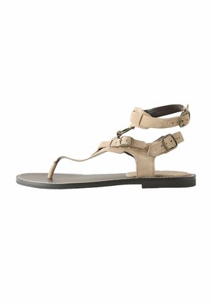 Sandalo piatto beige con suola nera, dotato di tre cinturini con fibbia regolabile intorno alla caviglia e al piede, e un anello per l'alluce.