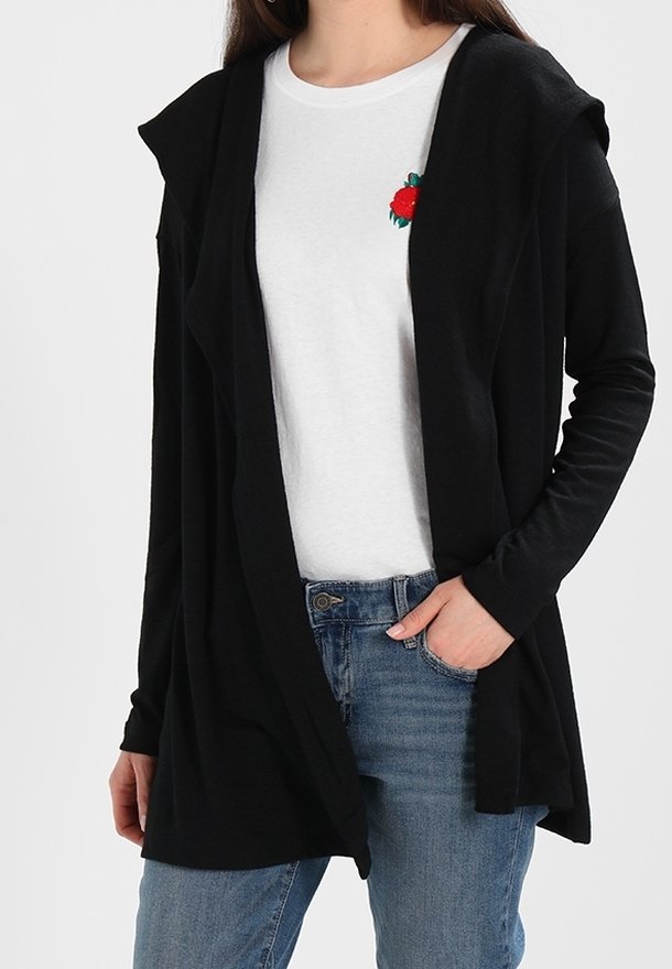 Femme portant un cardigan noir ouvert sur un t-shirt blanc avec un motif de rose rouge, associé à un jean bleu, main dans la poche.