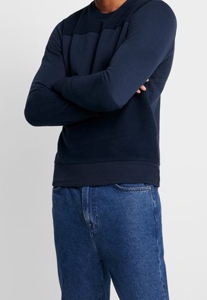 Mężczyzna ubrany w granatowy sweter z długim rękawem i niebieskie dżinsy, stojący z założonymi ramionami na jasnym, jednolitym tle.