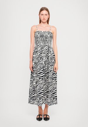 Maxi-jurk in zwart-wit zebra print, met spaghettibandjes, een gesmokt lijfje en een vloeiende rok met plooien. Zwarte platte schoenen.
