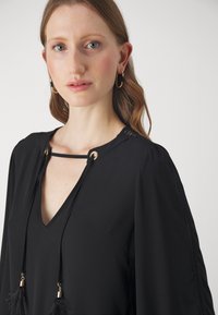 Black blouse with V-neck and front tie detail. Features gold hardware and delicate fringe accents on sleeves. Smooth, textured fabric.