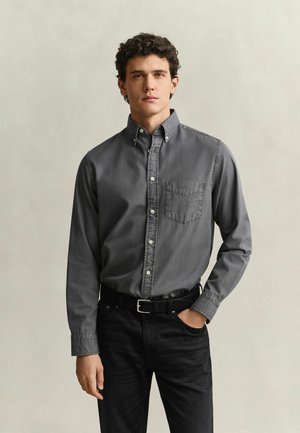 Jeune homme aux cheveux bouclés portant une chemise grise à manches longues boutonnée et un pantalon noir, debout avec une main dans la poche devant un fond uni.