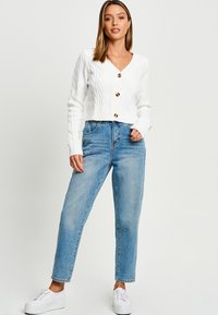 Cardigan court en tricot blanc avec un col en V et trois boutons marron, associé à un jean droit taille haute bleu clair et des baskets blanches.