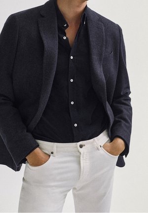 Blazer navy su una camicia nera, abbinato a pantaloni bianchi. Il blazer presenta un collo a lappola, mentre la camicia ha un collo button-down.
