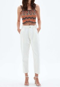Häkel-Crop-Top mit orangefarbenem, braunem und cremefarbenem Zickzackmuster. Hochtaillierte, weiße Hose mit lockerer Passform und gerafftem Taillenbund. Weiße Sandalen.