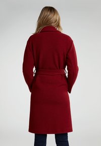 Manteau en laine rouge avec un large col et une taille ceinturée, doté de manches droites et atteignant la mi-mollet.