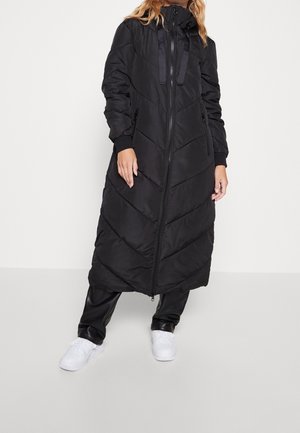 Frau in langem, schwarzem Steppmantel mit Reißverschluss, schwarzer Hose und weißen Sneakers, die vor einem schlichten hellen Hintergrund steht.