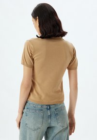 Haut en tricot beige à manches courtes avec une silhouette ajustée, des poignets et un ourlet côtelés. Porté avec un jean taille haute bleu clair. Texture lisse.