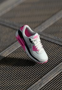 Nike sportbačiai, turintys baltą tinklelio viršutinę dalį su rožinėmis akcentais ir juodu padu. Dizainą papildo raišteliai ir firminis liežuvėlis.