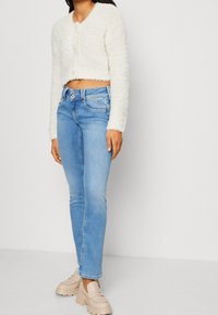 Žena v bílé huňaté zkrácené kardiganu, světle modrých vysokých džínách s dvojitým zapínáním a béžových silných mokasínech na podpatku.