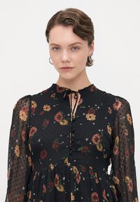Svart blomstret kjole med lange gjennomsiktige ermer, med prikkete tekstur, samlet liv og knytehals. Utsmykket med fargerike blomster og knapper.