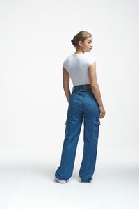Young woman with hair clip wears white fitted t-shirt and high-waisted blue cargo jeans, standing and looking to the side on white background.